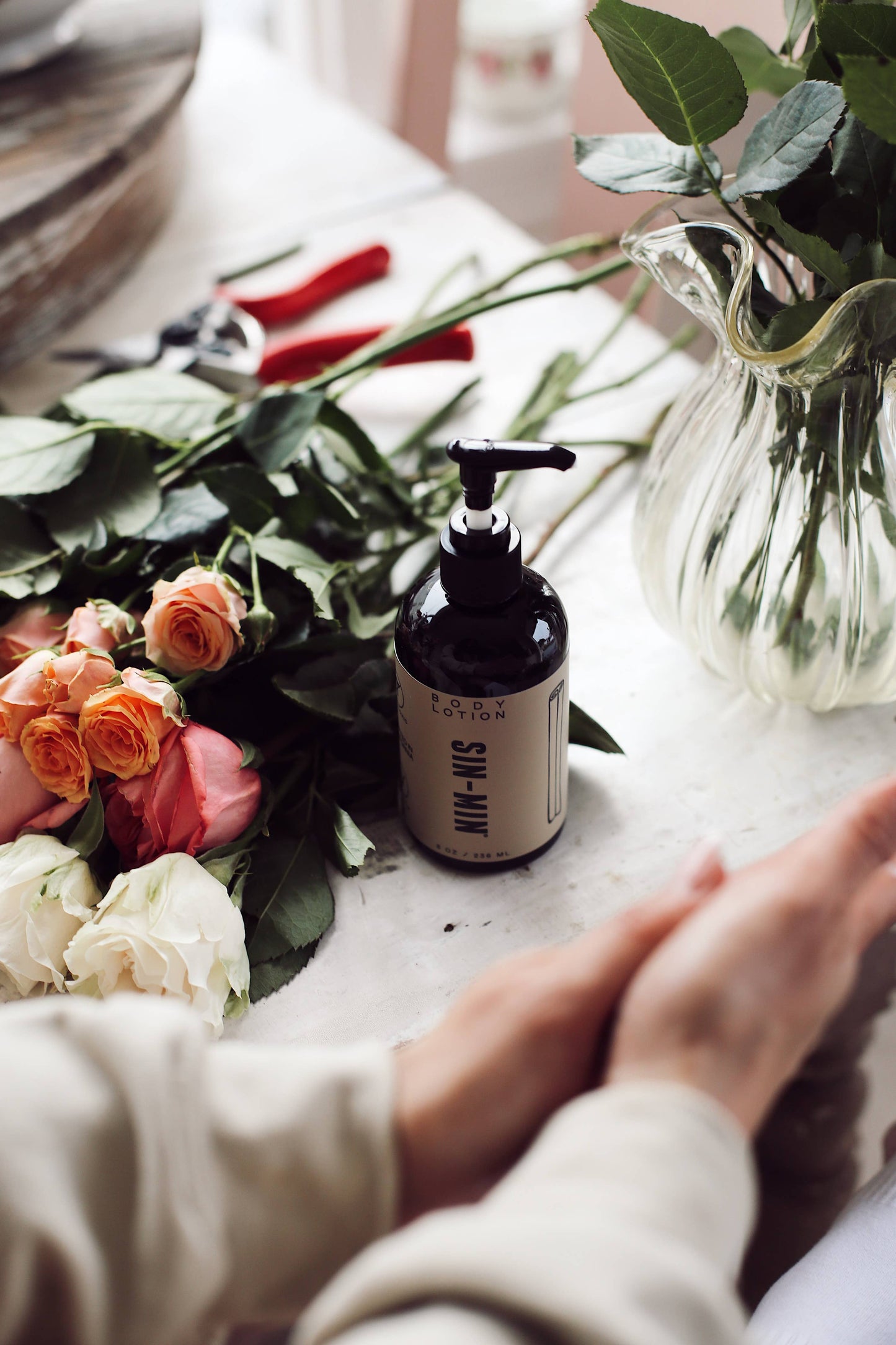 SIN-MIN sweet cinnamon vanilla body lotion bottle with pump near fresh roses and hands on marble table