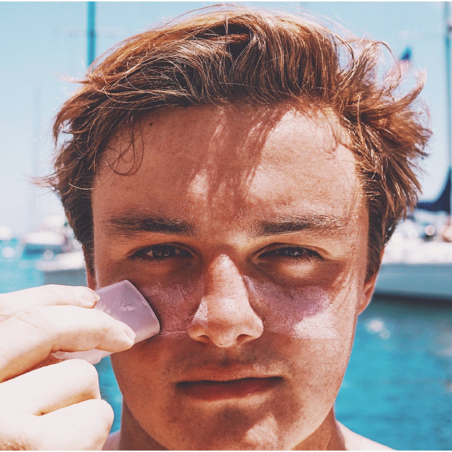 Young man applying indie sunscreen stick on his cheek outdoors near water with boats in background