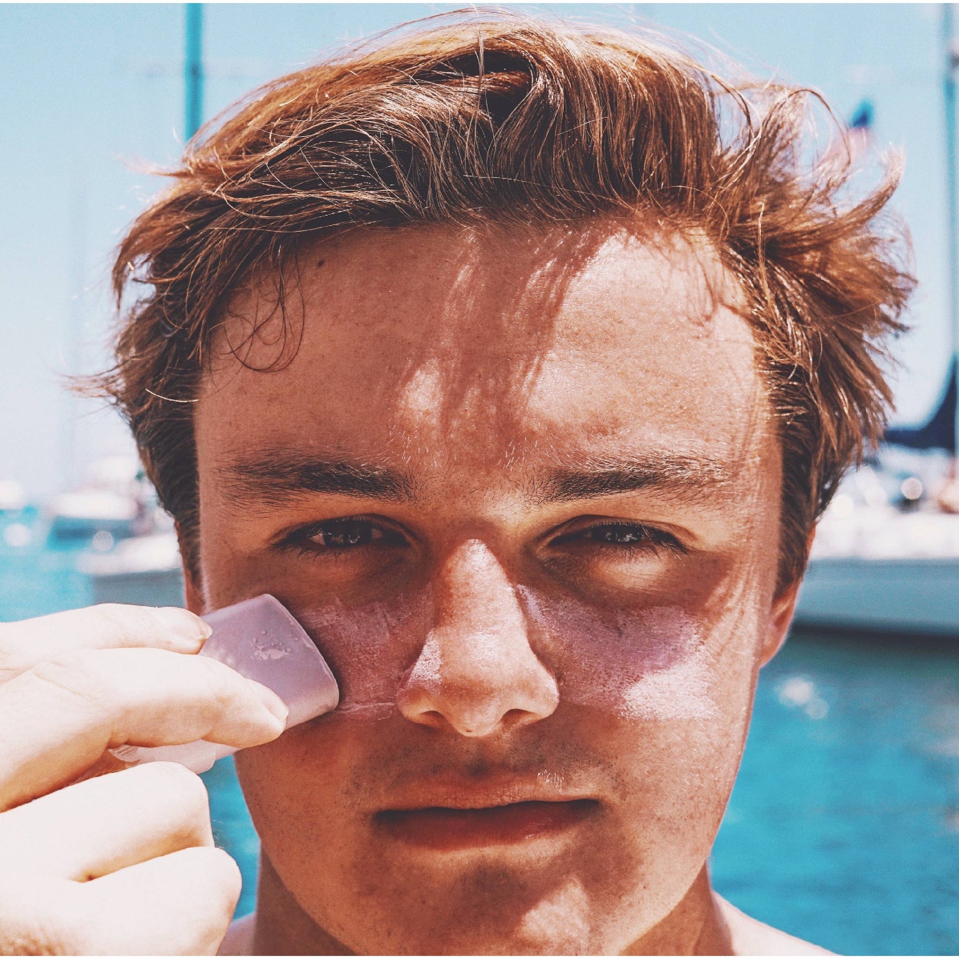 Young man applying indie sunscreen stick on his cheek outdoors near water with boats in background