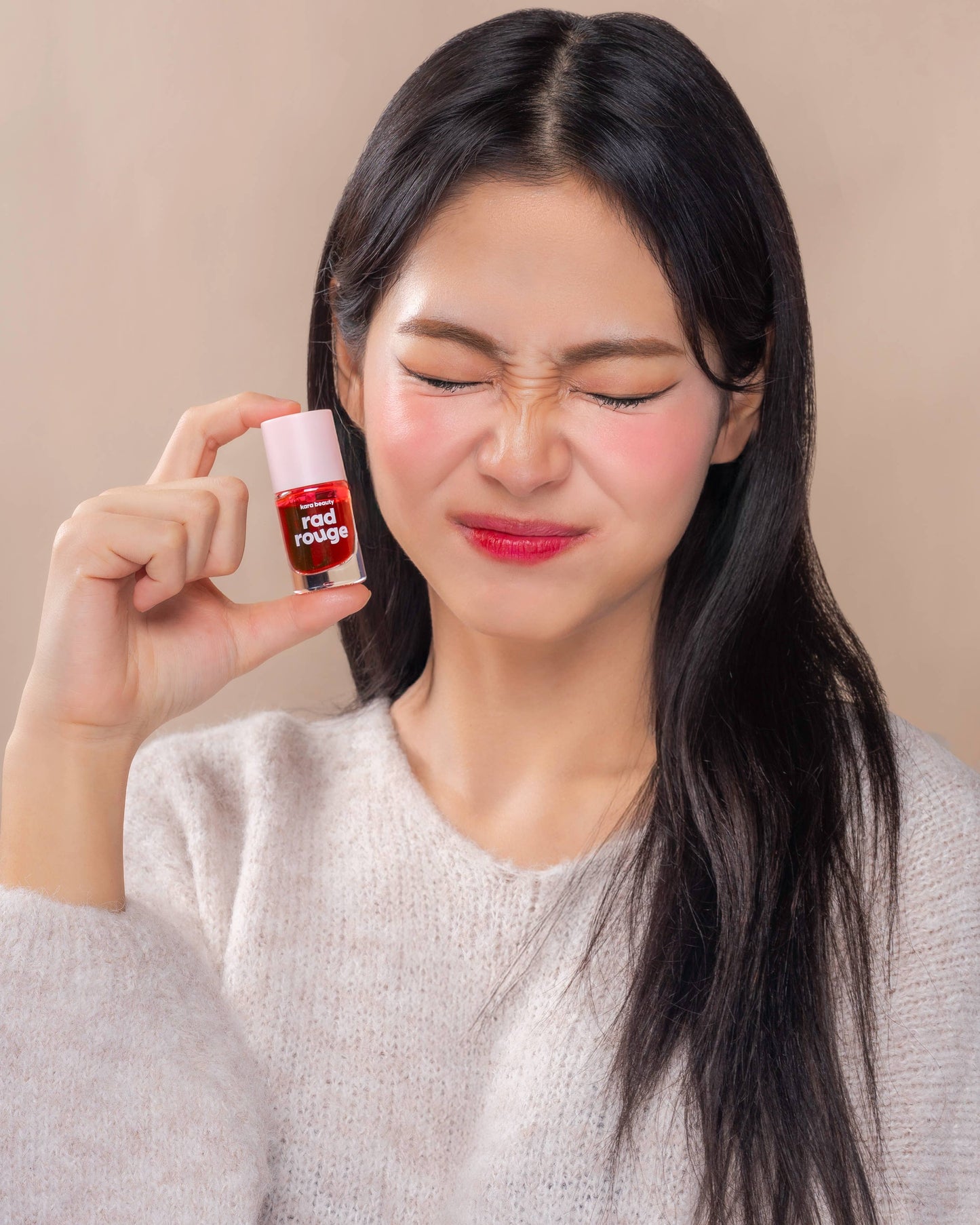 Young woman scrunching eyes and smiling while holding Kara Beauty Rad Rouge Lip Tint near her face