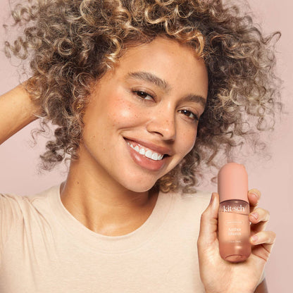 Smiling woman with curly hair holding a bottle of Innovate the Label amber hair perfume