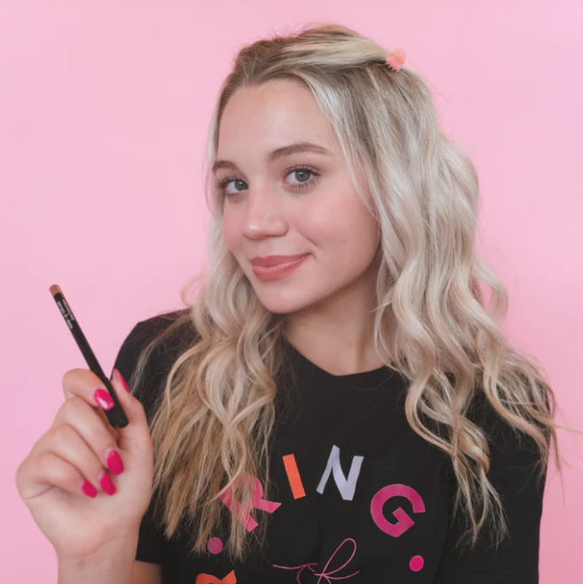 Woman holding creamy lip liner crayon by Innovate the Label Beauty Bundles with wavy blonde hair and pink background