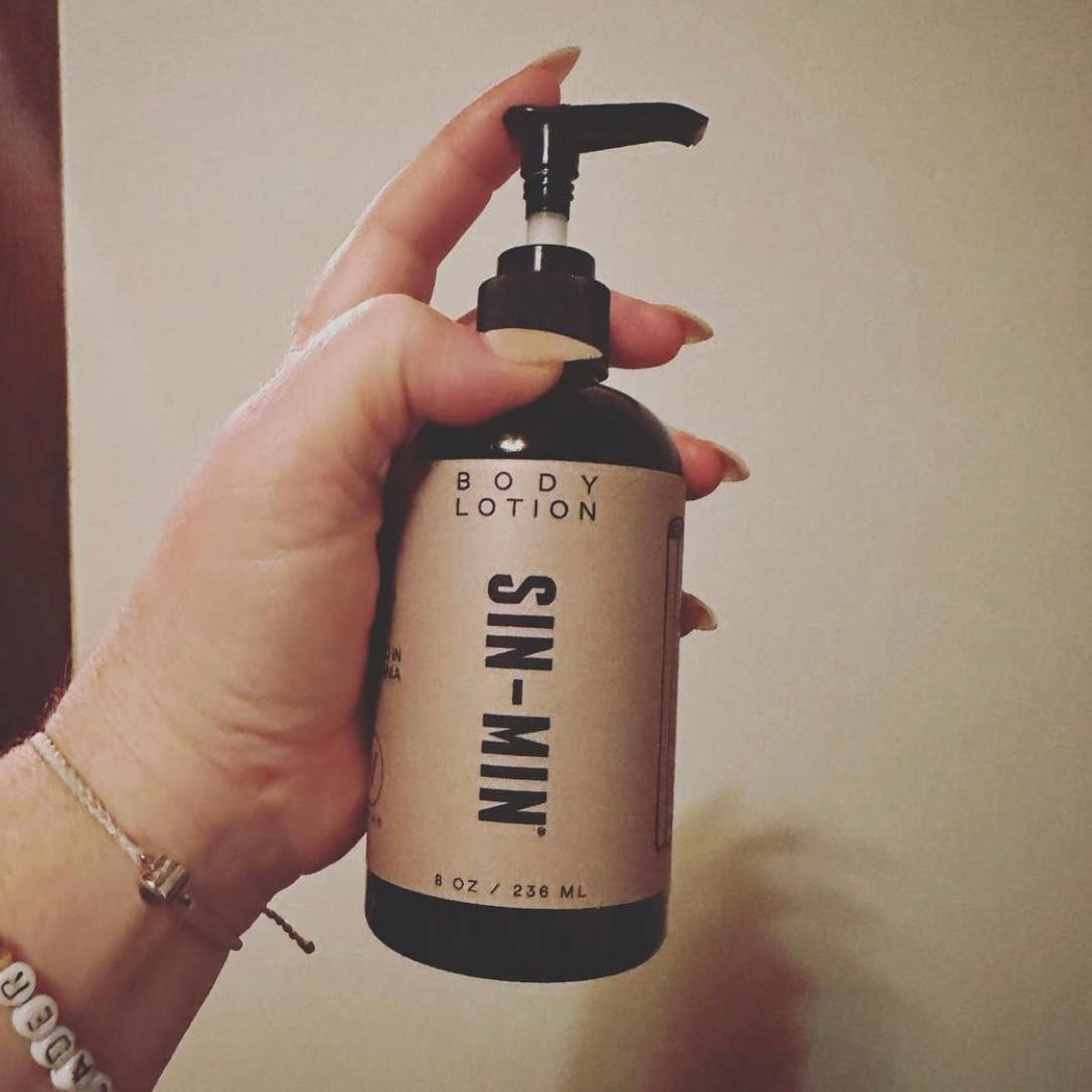 SIN-MIN sweet cinnamon vanilla body lotion in a pump bottle held by a hand with neutral background