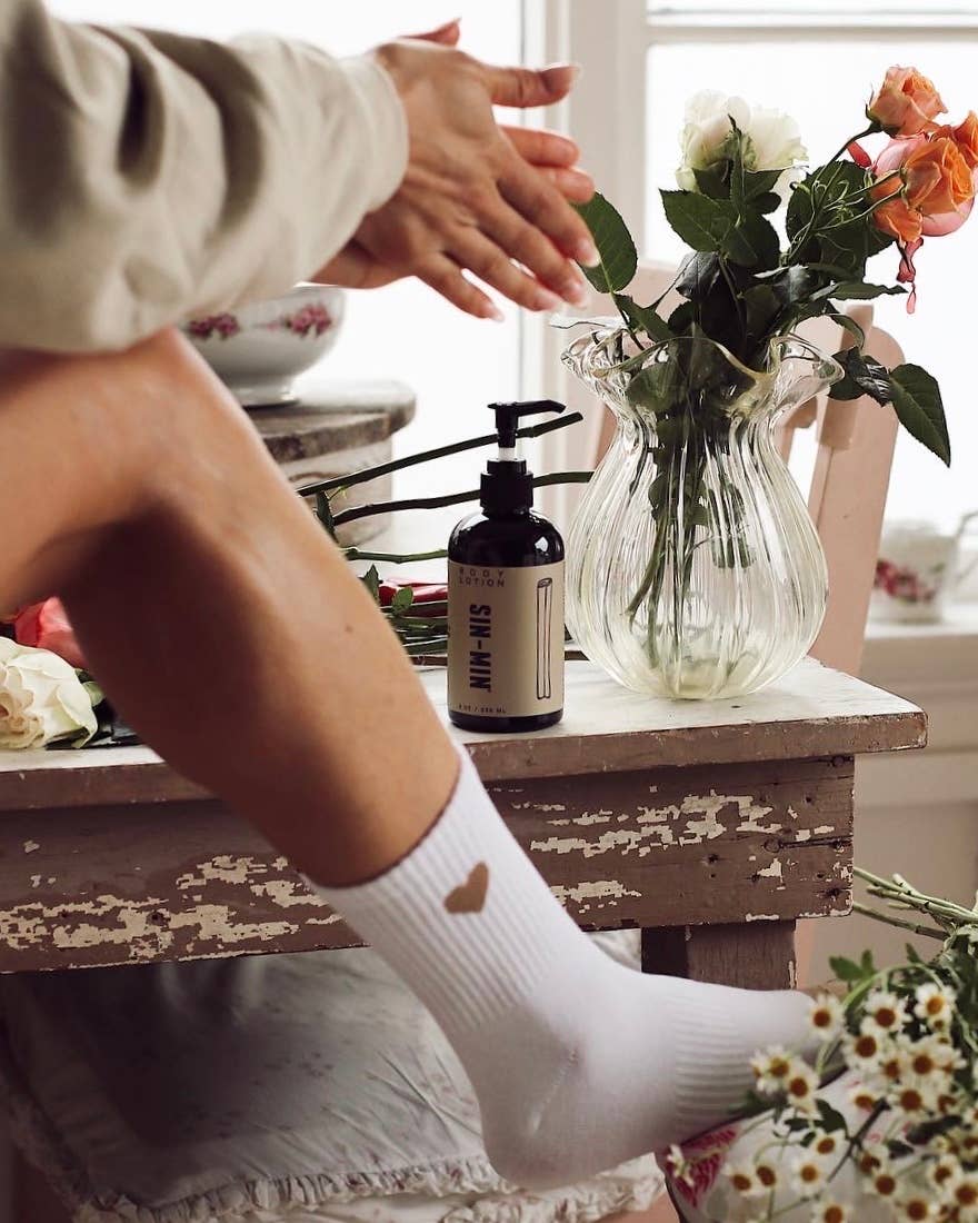 SIN-MIN sweet cinnamon vanilla body lotion bottle on rustic table near flowers and person applying lotion