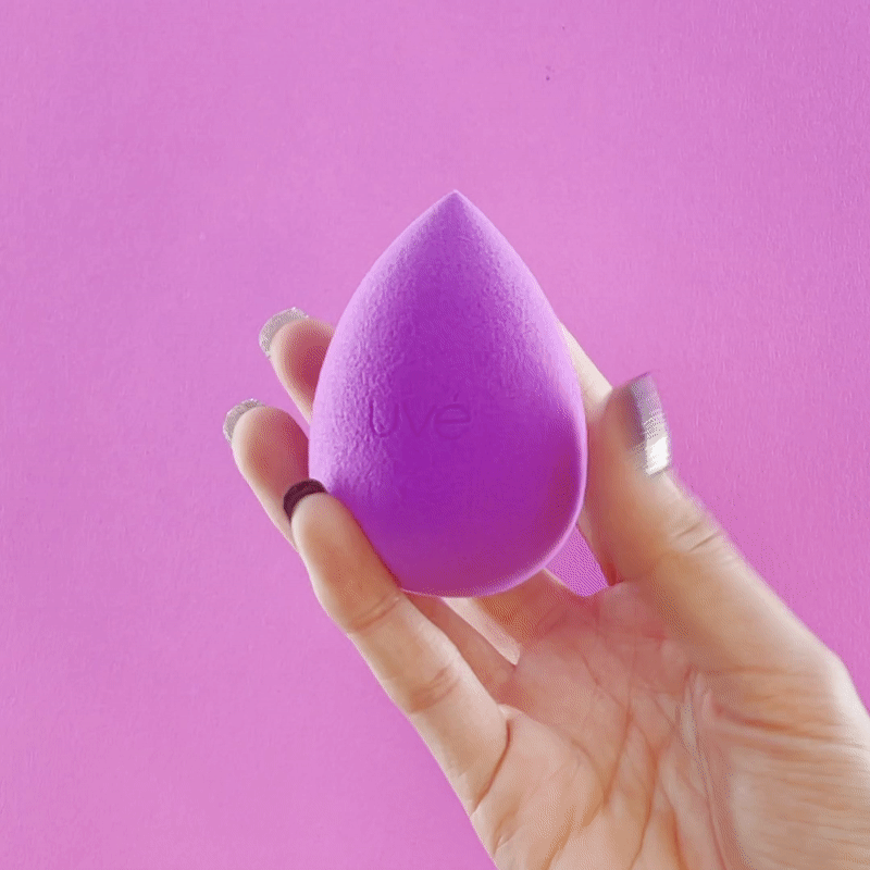 Innovate the Label ultra soft makeup sponge in purple held by hand against pink background